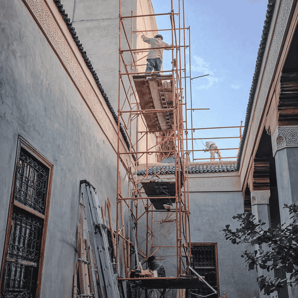 Rénovation et travaux de construction d’un riad à Marrakech Ouvriers marocains en chantier dans un riad en rénovation à Marrakech, avec échafaudages, sacs de matériaux, arches anciennes et patio en cours de réhabilitation.